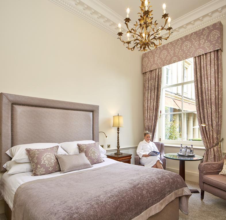 Macdonald Bath Spa Hotel Ngoại thất bức ảnh The photo shows a well-appointed hotel room that features a large, elegant bed with soft bedding and decorative pillows. The walls are painted a light color, and there is an ornate chandelier hanging from the ceiling. A beautiful window is adorned wi