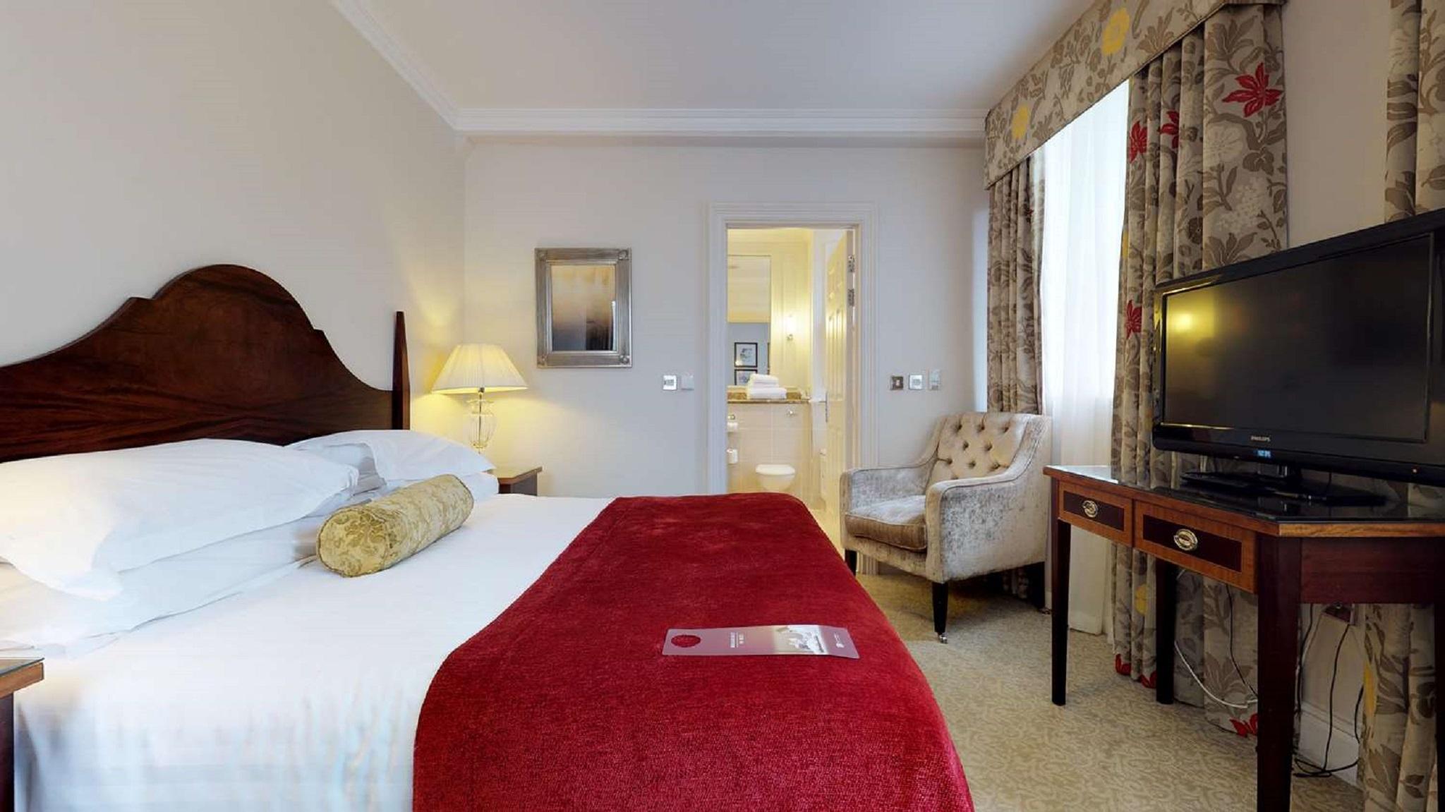 Macdonald Bath Spa Hotel Ngoại thất bức ảnh The photo shows a hotel room with a queen-sized bed covered in white linens and a deep red throw blanket. There is a decorative pillow on the bed. Next to the bed, there's a small nightstand with a lamp. In the background, a seating area features a b